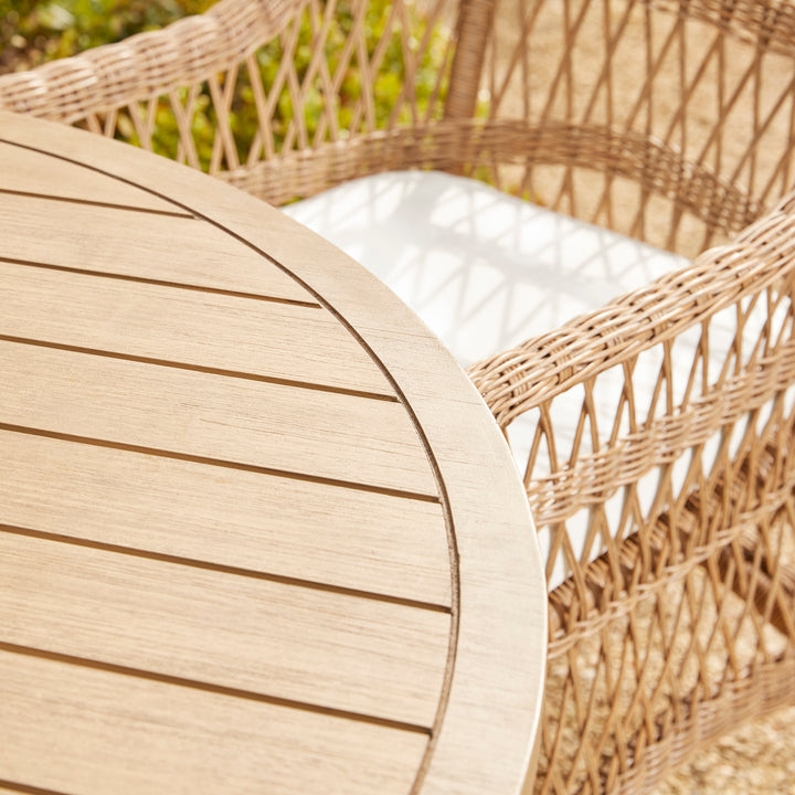 A solid acacia wood garden dining table and woven armchair with white seat cushion.