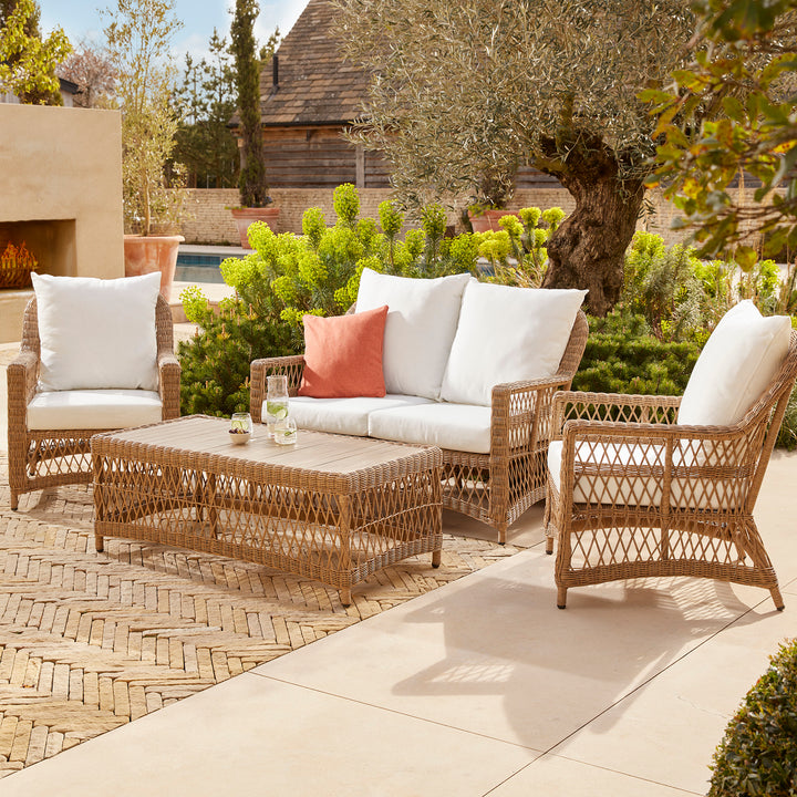 Stylish outdoor wicker furniture set featuring a four-seater sofa, two armchairs, and a matching coffee table with white cushions and rust-toned accent pillows, beautifully arranged on a sunlit patio surrounded by lush greenery and a modern stone fireplace.