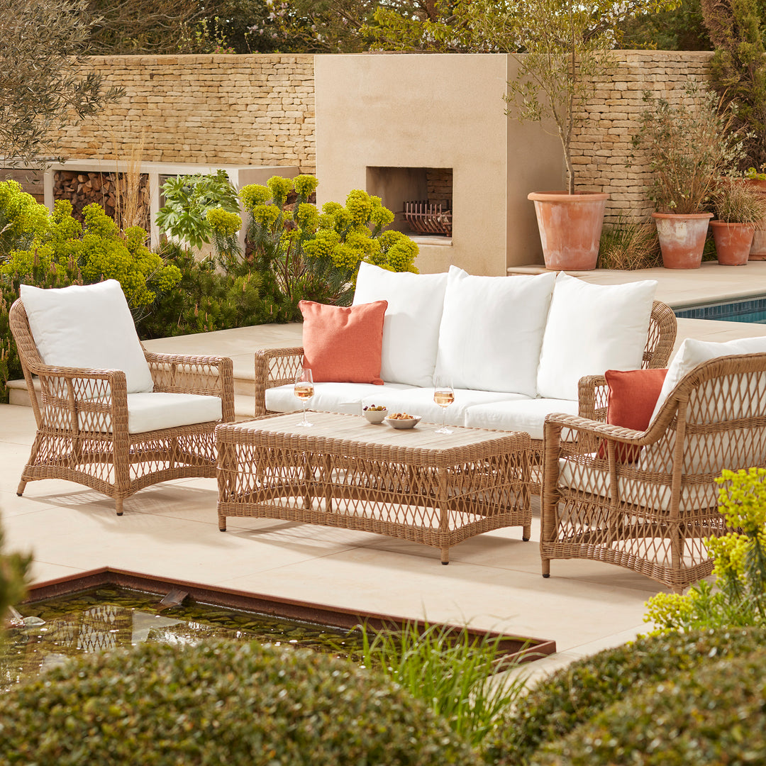 Stylish outdoor wicker furniture set featuring a five-seater sofa, two armchairs, and a matching coffee table with white cushions and rust-toned accent pillows, beautifully arranged on a sunlit patio surrounded by lush greenery and a modern stone fireplace.