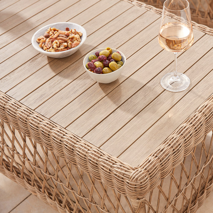 Close-up of a wicker coffee table with a natural wood top, featuring bowls of mixed nuts and olives alongside a glass of rosé wine, perfectly styled for relaxed outdoor dining.