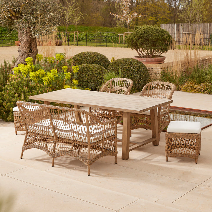 Solid acacia wooden garden table with two stools, two dining chairs and a casual dining sofa, styled on a stone terrace amidst sculpted greenery.
