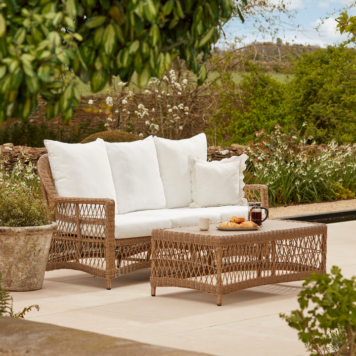 Outdoor wicker sofa and coffee table set with plush white cushions, styled on a sunlit patio, surrounded by lush garden greenery, offering a timeless and refined alfresco living experience.