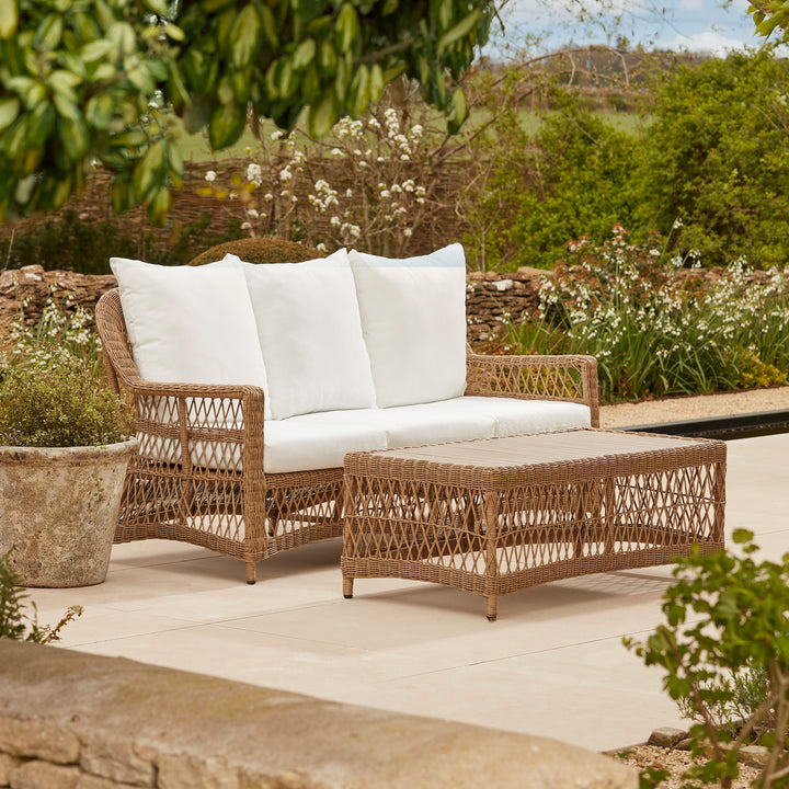 Solid acacia wooden garden table in an open weave rattan frame, with deep comfortable sofa, styled on a stone terrace amidst sculpted greenery.