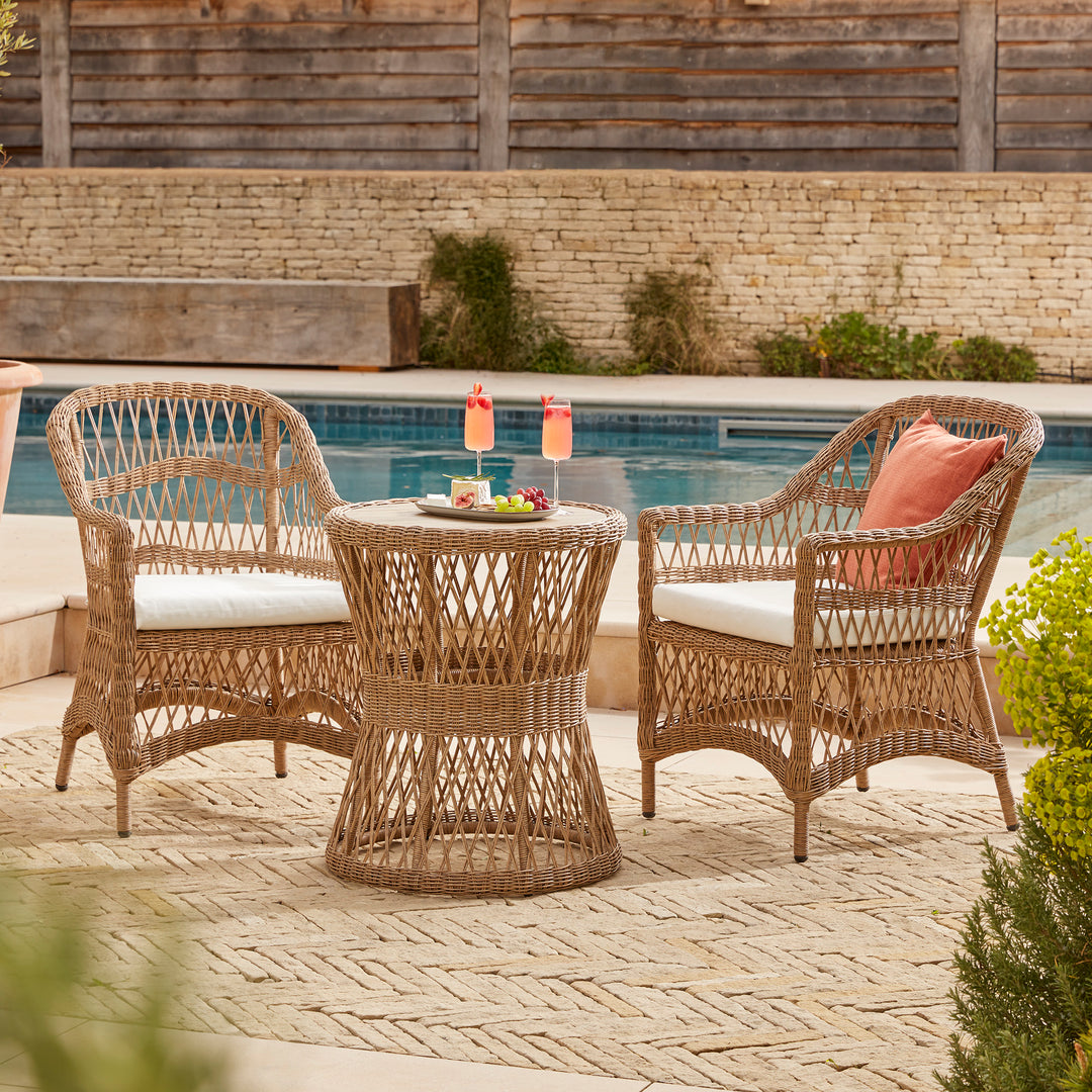 A two-seater round bistro table and chair set featuring a wooden and open weave table and wicker-style armchairs. Set on a cotswolds chevron brick patio