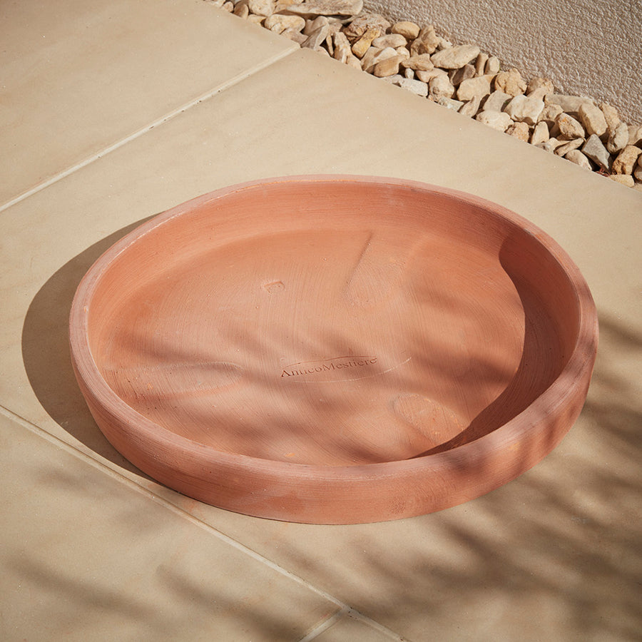 An Italian terracotta plant pot saucer placed on a buff coloured patio with the dappled sunlight shining through an olive tree onto the floor. A timeless terracotta saucer to protect your patio and house your italian garden pots indoors or outdoors.