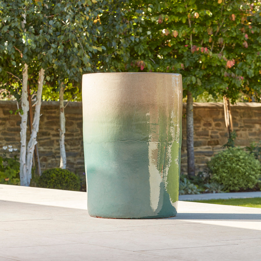 Tall ceramic garden pot with soft turquoise and cream reactive glaze, positioned in dappled sunlight on a stone patio.