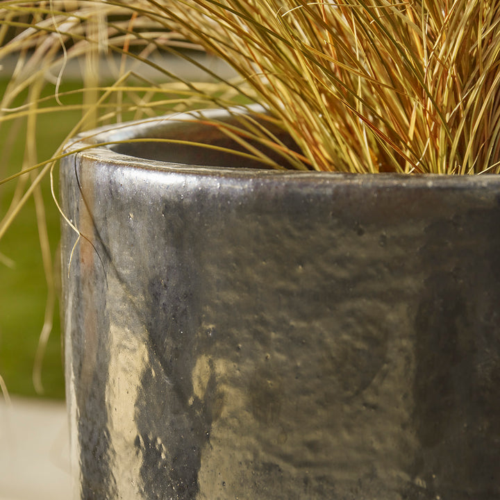 Detail of tall black ceramic plant pot with hand-glazed glossy finish, shown with ornamental golden grass planting on a patio.