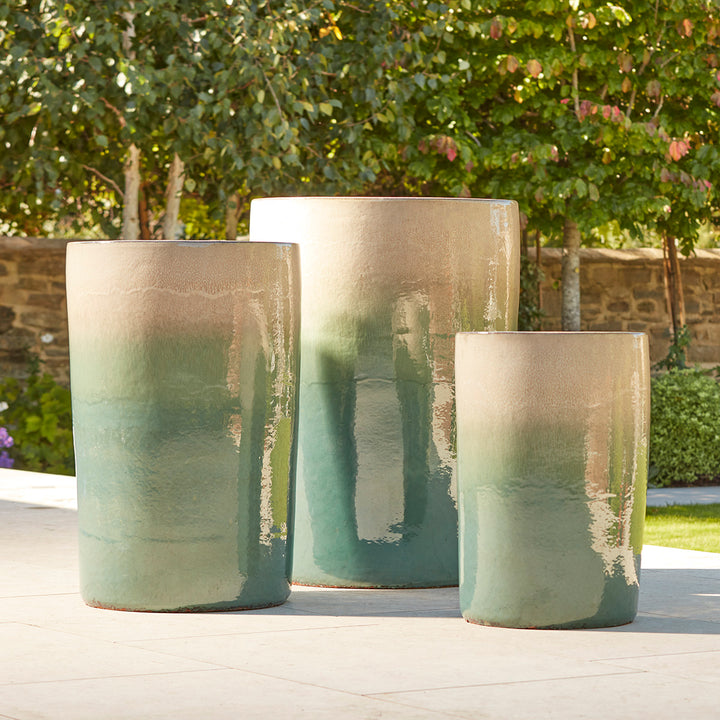 Trio of tall cylindrical ceramic pots with reactive turquoise and cream glaze, displayed outdoors in a formal garden setting