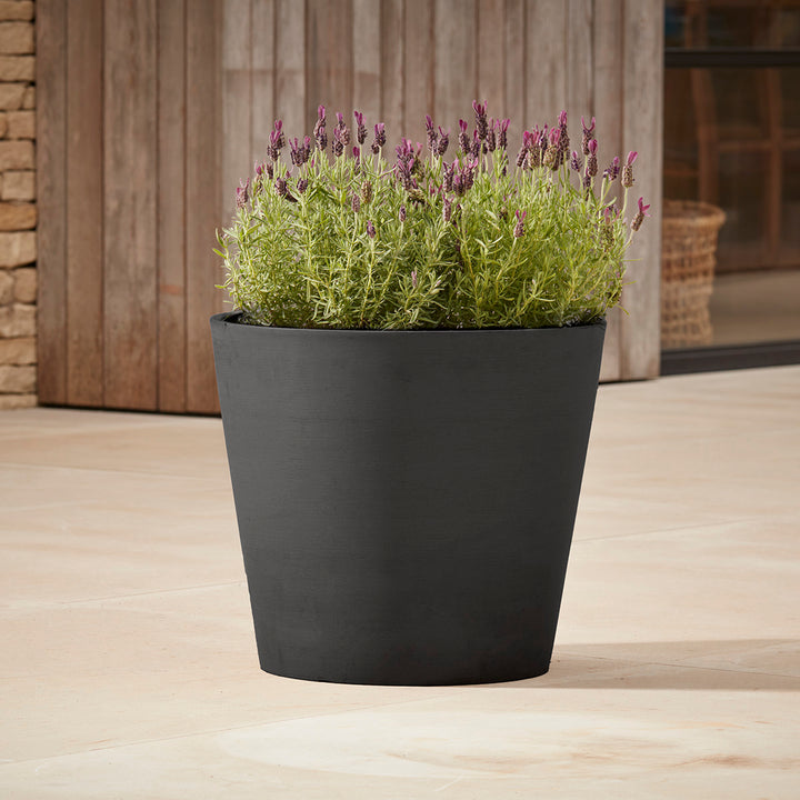 Gardenesque x EcoPots Round Plant Pot in charcoal, featuring a sleek conical shape, placed on a patio with lavender plants.