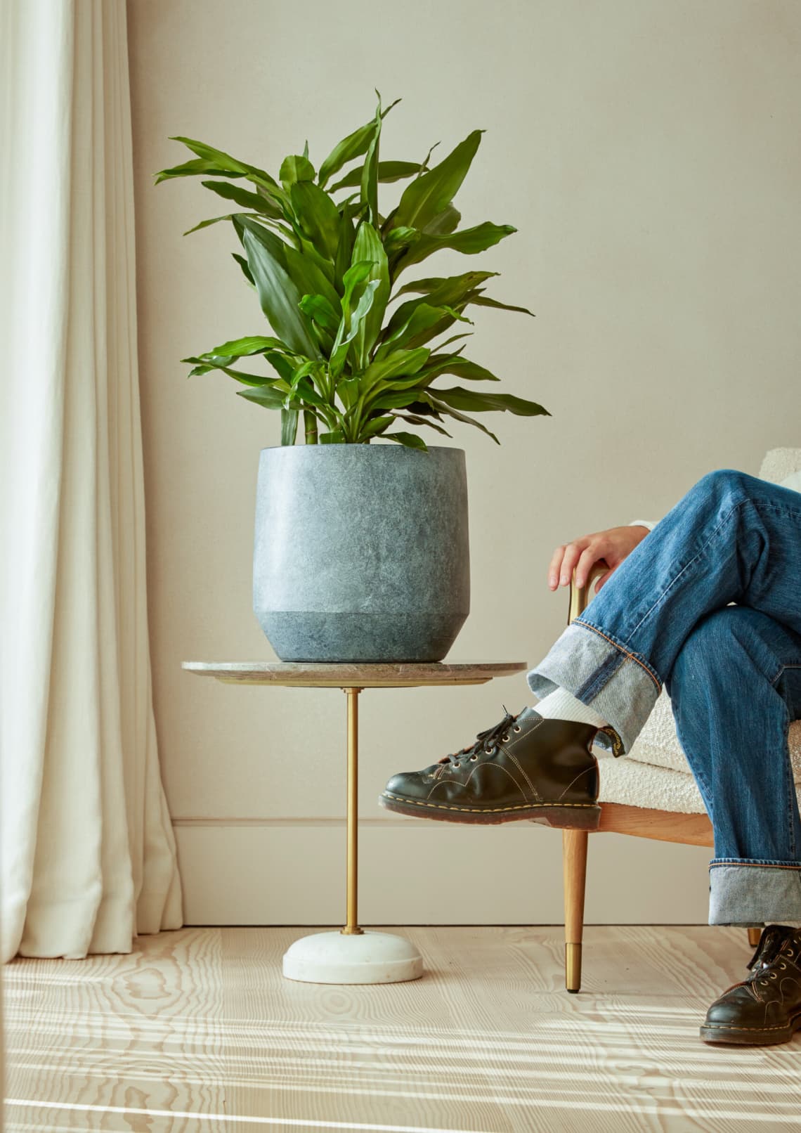 A modern interior scene featuring a green leafy plant in a two-toned gray planter, sitting on a round side table with a brass stand and a marble base. The background includes a neutral-toned wall and floor-length cream curtains. To the right, a person is partially visible, sitting on a mid-century modern wooden armchair, wearing cuffed blue jeans, white socks, and black leather boots, with one foot resting on the table's edge.