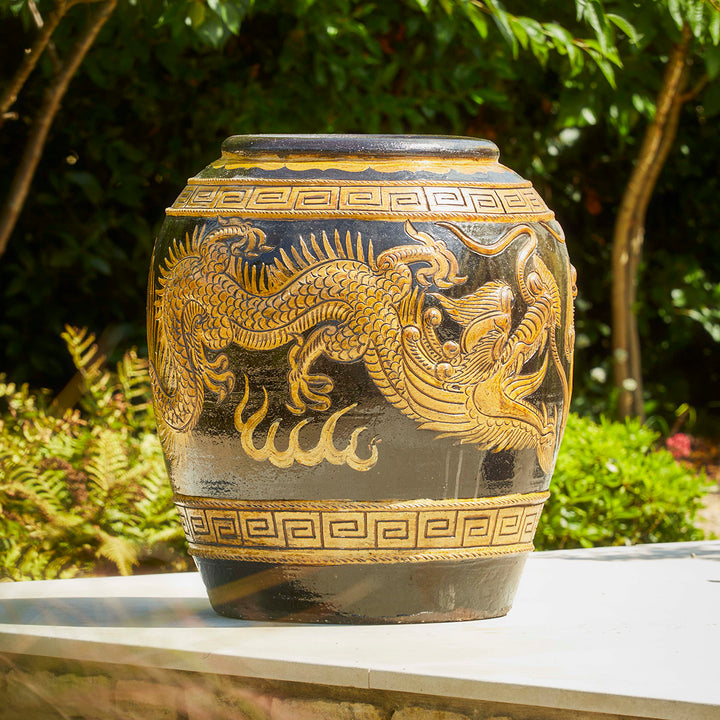 Large antique-style Chinese garden pot with detailed dragon motif, placed outdoors on a stone surface surrounded by greenery.