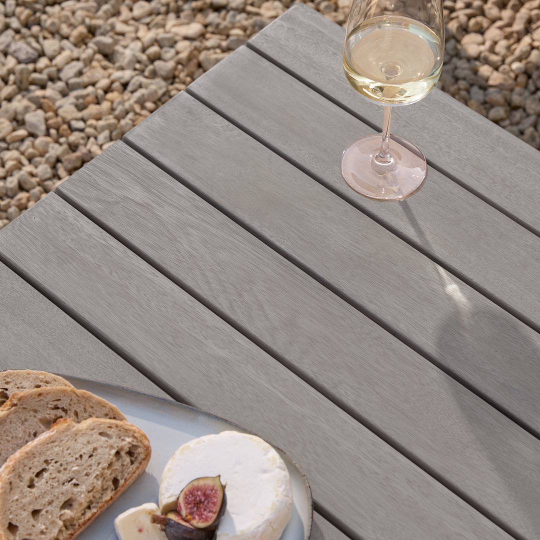 Repton Classic Wooden Garden Coffee Table with a slatted design, part of the Repton Corner Garden Furniture Set. Styled on a stone patio with two glasses of white wine and a bowl of snacks, this weather-resistant outdoor table complements modern patio and garden seating areas.