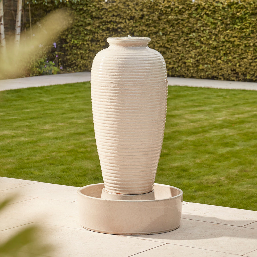 Tall cream glazed ripple urn water feature in garden patio setting, with cascading water over textured ribbed ceramic urn and round basin.