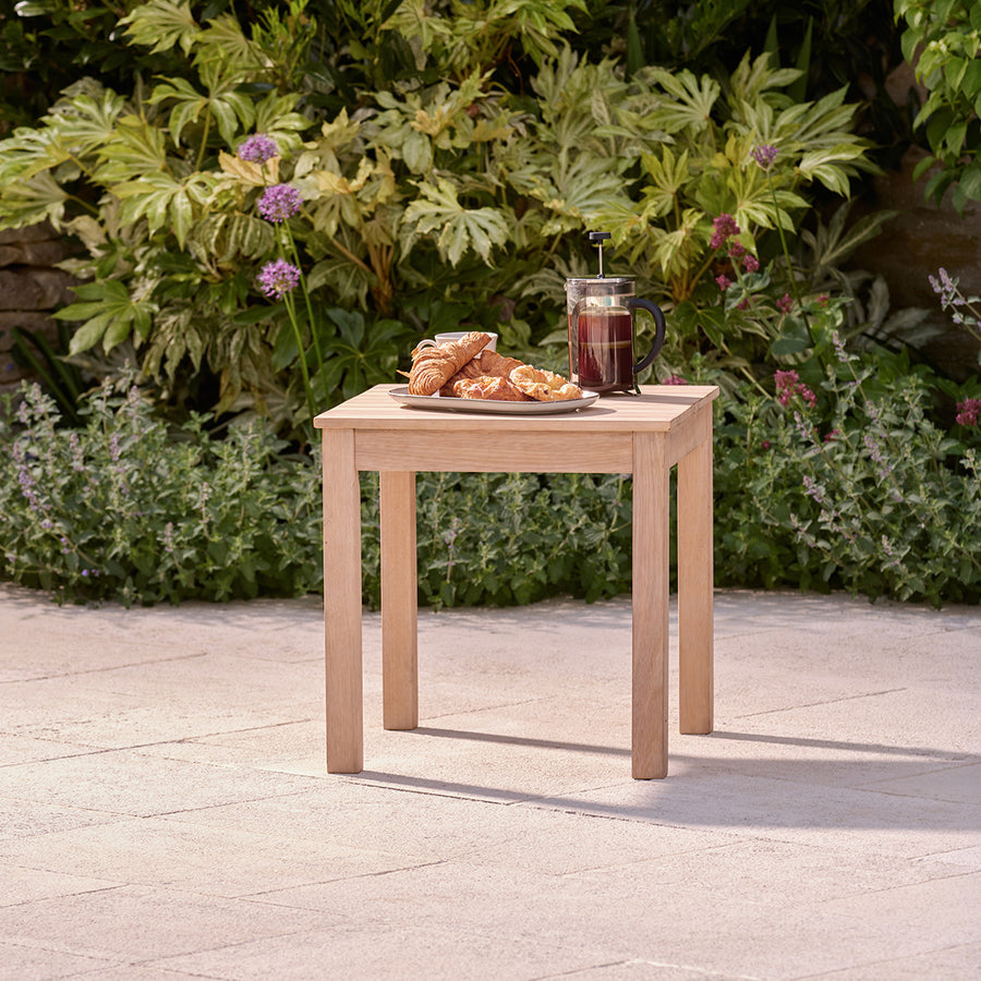 Wooden garden furniture side table styled with pastries and coffee on a patio surrounded by planting.