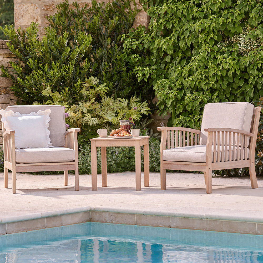 Wooden garden furniture table with two armchairs by a pool, styled for outdoor breakfast with natural cushions.
