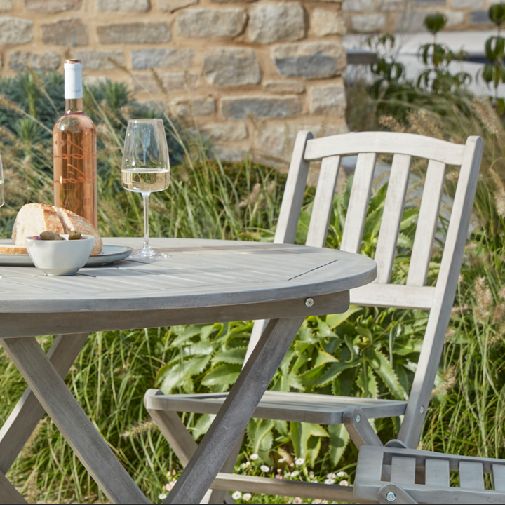 Close-up of the Repton Wooden Dining Set, featuring a round foldable table and a matching wooden chair with a slatted backrest in a weathered grey finish. The table is set with a bottle of wine, a glass, and a small plate with bread and olives. Positioned on a stone patio with a rustic brick wall and greenery in the background, designed for outdoor dining and garden spaces.
