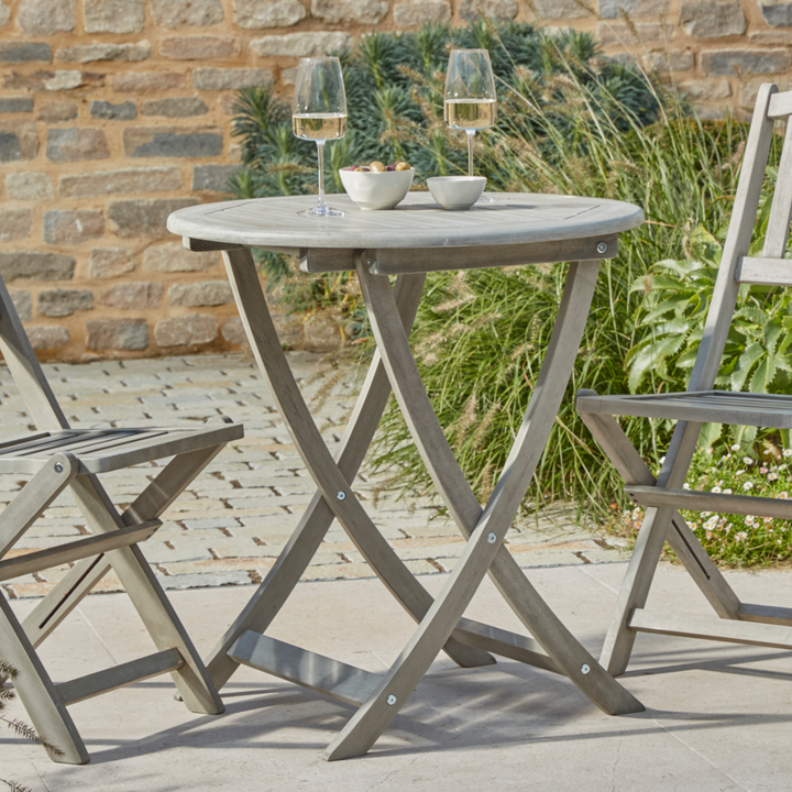 Close-up of the Repton Wooden Bistro Set, showcasing a round, foldable wooden table with a weathered grey finish. The outdoor dining setup features two wine glasses and small snack bowls, positioned against a rustic brick wall and lush garden greenery. Perfect for stylish and functional patio seating.