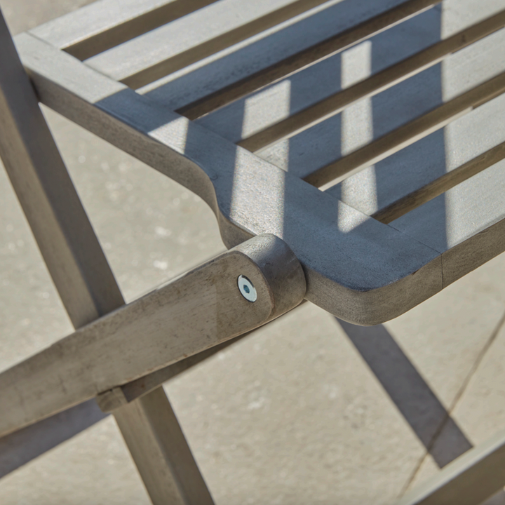 "Detailed close-up of the Repton Wooden Bistro Chair, showcasing its slatted seat, smooth weathered grey finish, and sturdy foldable frame. Sunlight casts soft shadows across the natural wood grain, highlighting the craftsmanship and durability of this outdoor garden chair, perfect for patios and balconies.