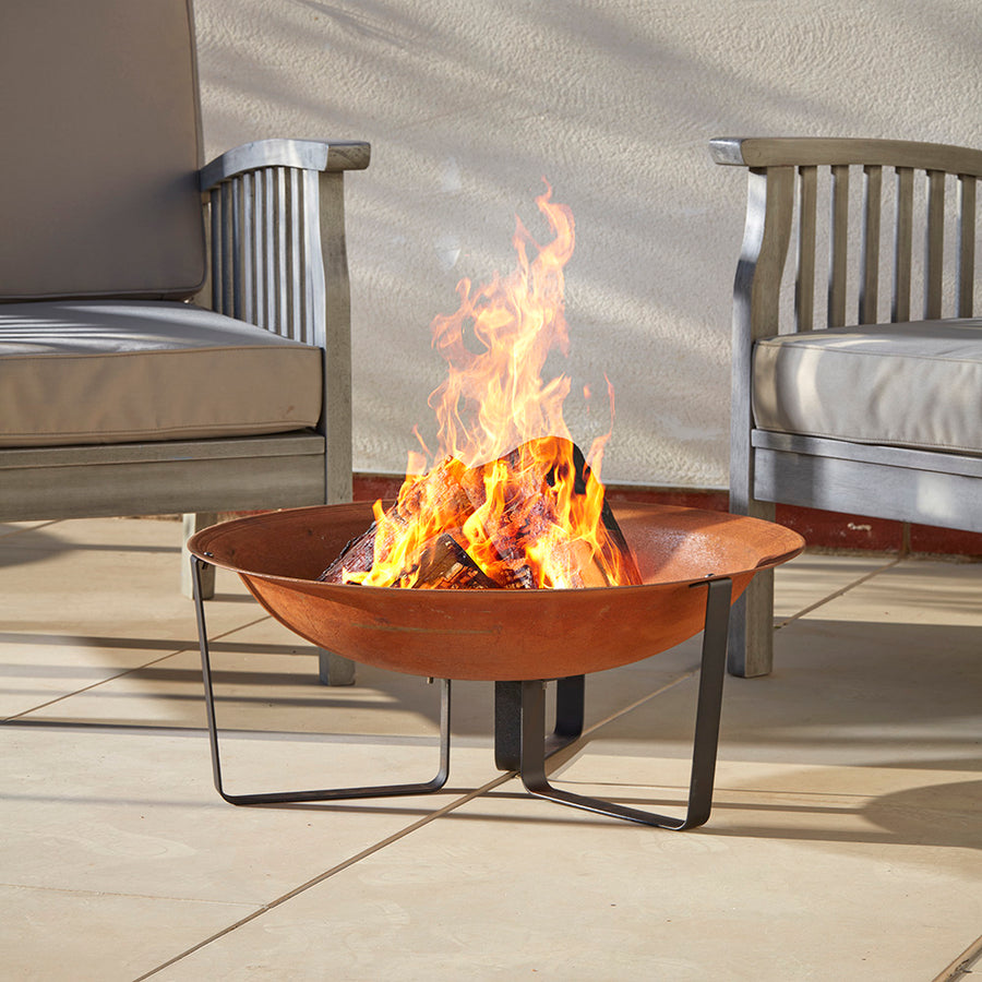 A rust-finish steel fire pit bowl with contemporary steel legs placed on a cotswold patio. The fire is lit inviting a cosy place to gather in the garden