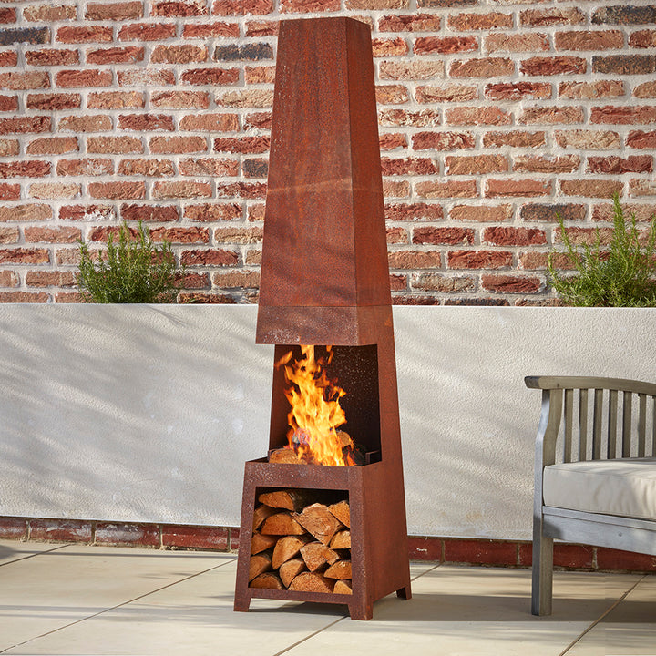 A tall rustic chimenea is standing on a cotswold courtyard garden on a spring evening. The bottom log storage is filled with kiln dried logs. In the top section, a fire is smoldering, sending the smoke up the chimenea so it's guests can enjoy the warmth