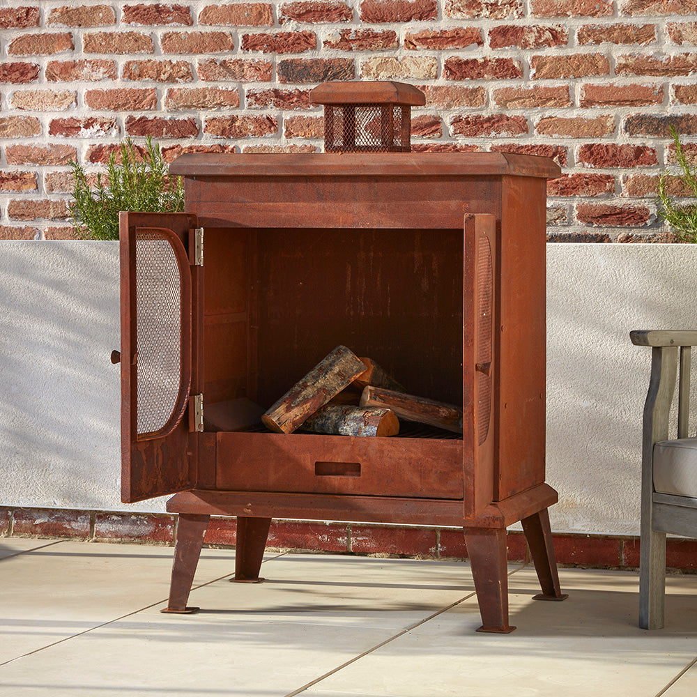 A Rustic garden wood burner set on a cotswold patio. The mesh doors are open and the the logs are stacked inside ready to be light.
