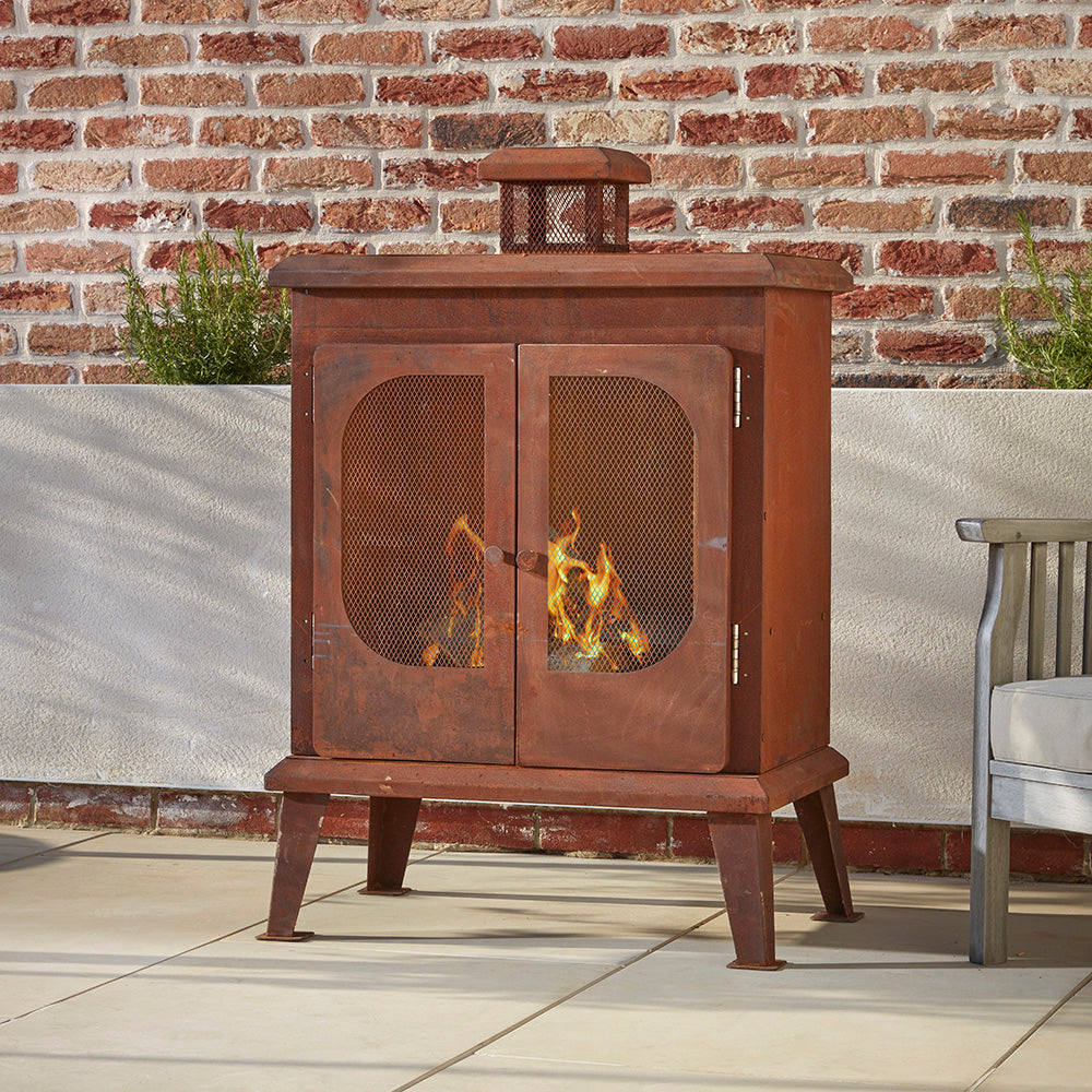 Wakehurst Outdoor Wood Burning Fireplace with a vintage rust finish, placed in a cozy garden setting. A freestanding steel log burner with mesh doors and a built-in chimney for efficient smoke control.