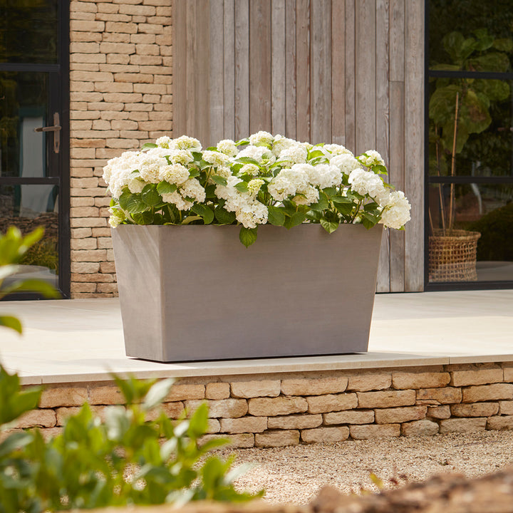 Gardenesque x EcoPots Extra Large Wheeled Trough in taupe, elegantly displayed with white hydrangeas on a patio.