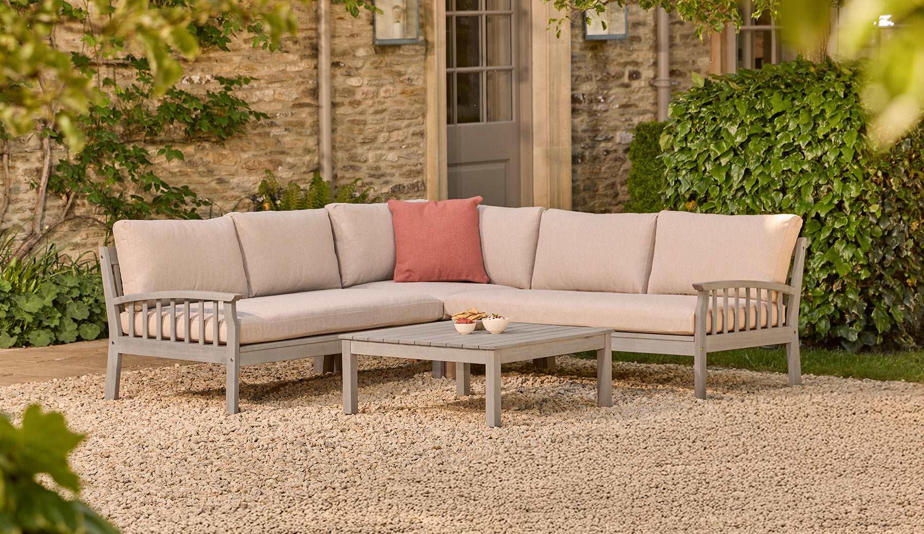 Outdoor wooden corner patio furniture set with beige cushions and a red pillow on a stone patio.
