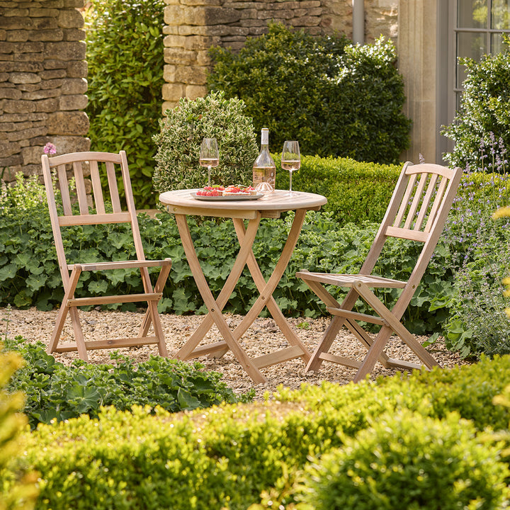 Wooden outdoor table with two chairs and a bottle of wine in a garden setting.