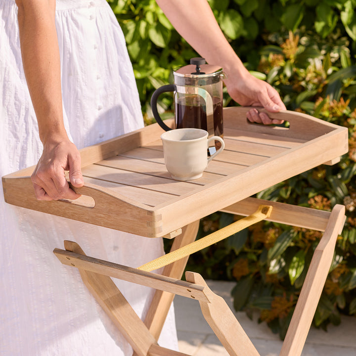 Person holding a wooden folding table with a cup and French press outdoors.