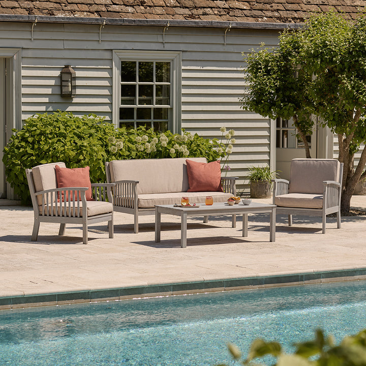 The Repton Wooden Lounge Set, featuring a two-seater sofa, two armchairs, and a matching coffee table in a weathered grey finish. The outdoor seating set includes comfortable beige cushions and is arranged on a stone patio against a rustic brick wall with lush greenery. Designed for stylish and relaxing garden lounging.