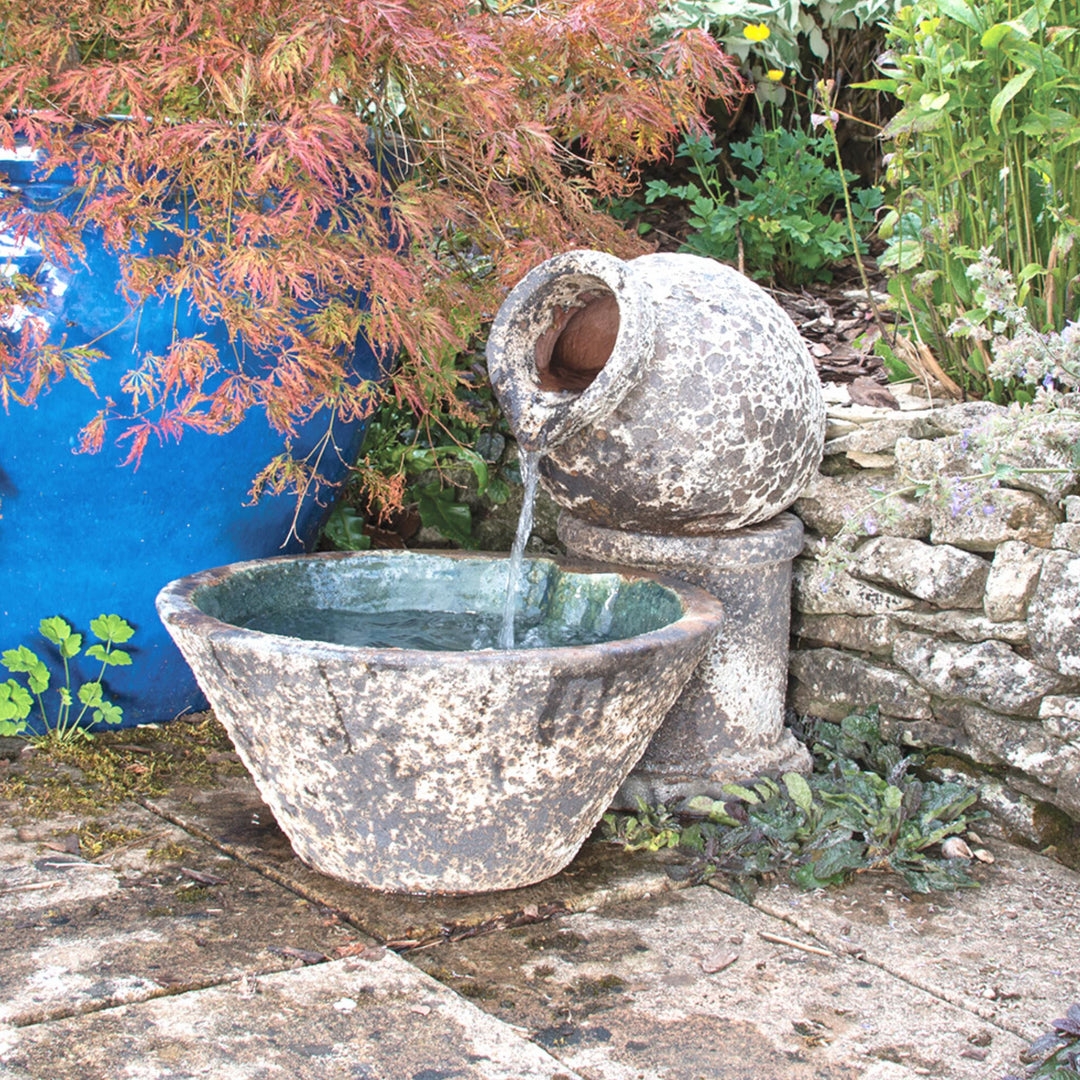A jug and bowl stone water feature with a rustic salt glaze finish, placed in a tranquil garden setting against a stone wall, with water flowing gently into the basin.