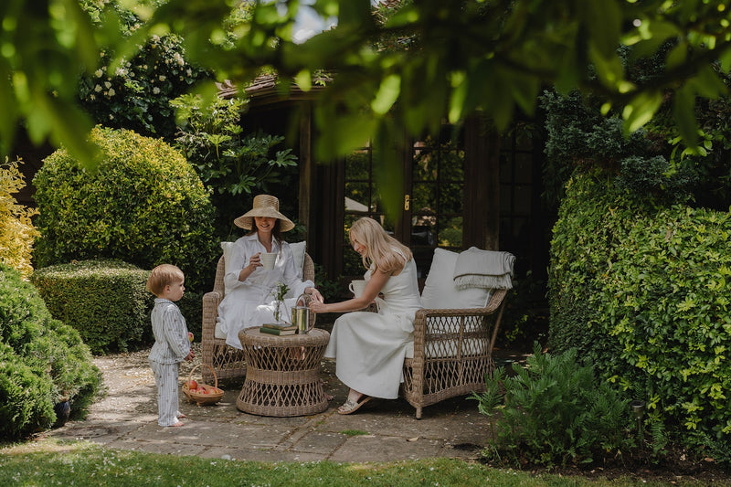 woven garden armchair and bistro table set in a small patio space under the dappled light of trees. 2 friends enjoying a quiet drink together 