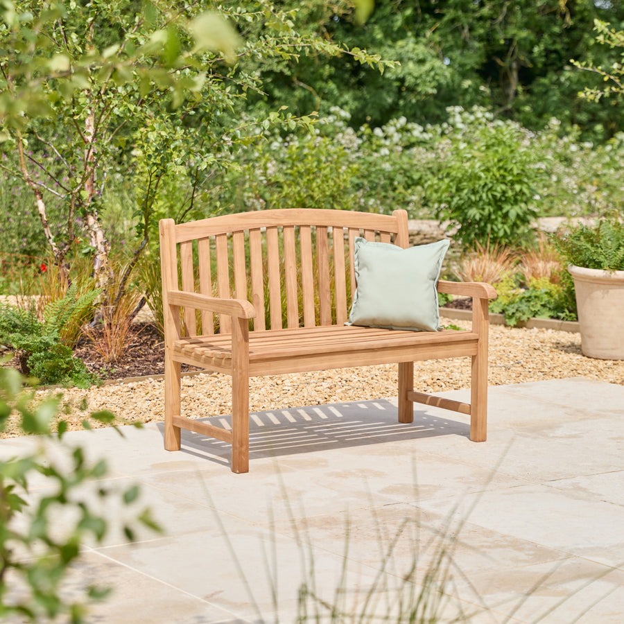 A wooden garden bench with a curved backrest and armrests, shown on a stone patio in a landscaped garden. A light sage green throw pillow rests on the bench.