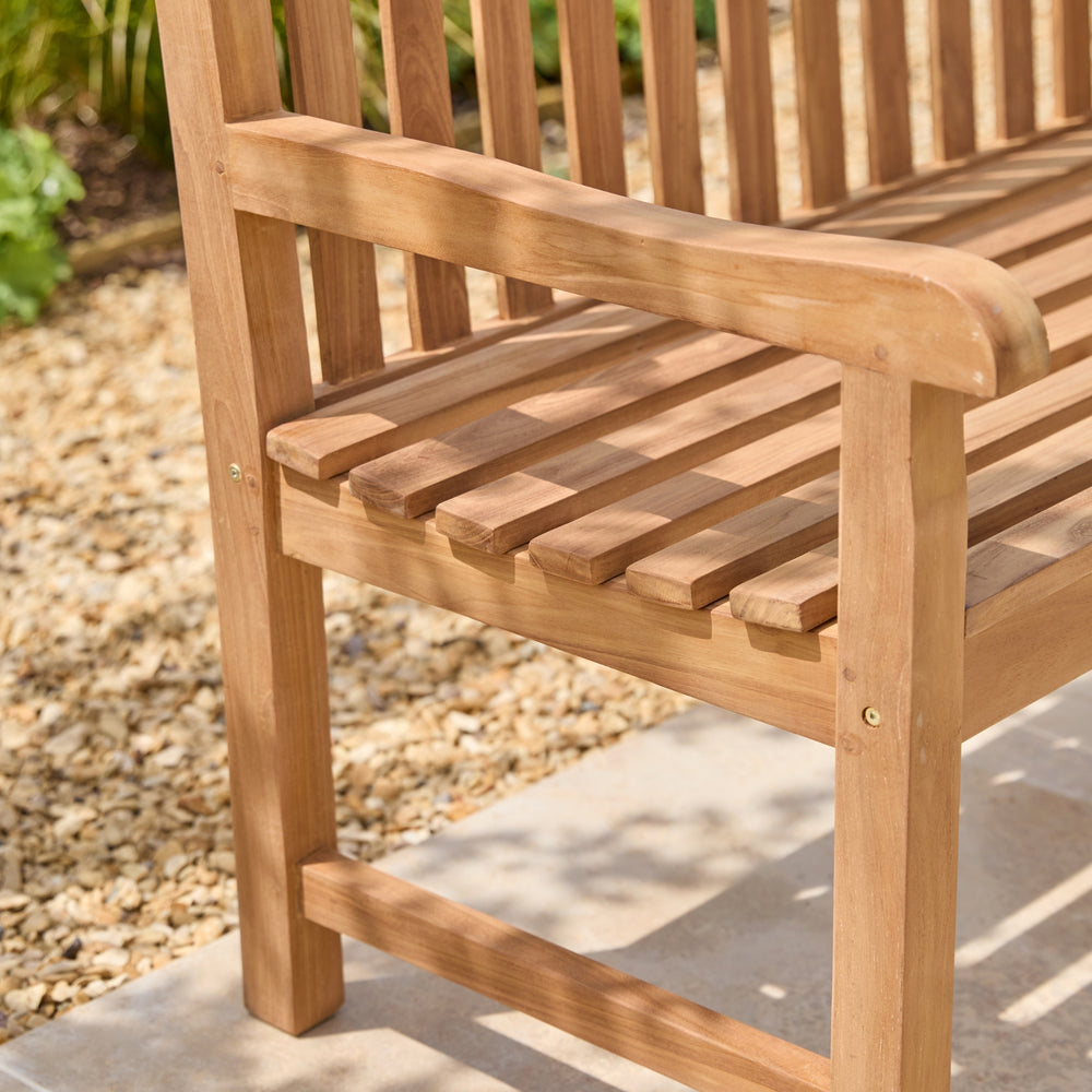 Close-up of a wooden slatted bench in a garden setting, showing the armrest, seat slats, and leg on a patio with gravel and plants in the background.