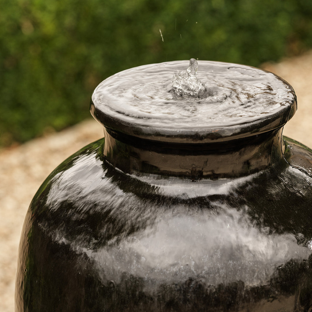 Detail of water flowing from the top of black urn water feature, smooth glazed finish reflecting sunlight.