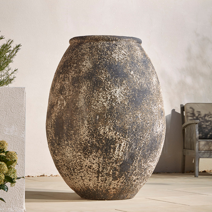 Extra Large 125cm tall stone antique effect jar  for plants. Positioned on a sun drenched Mediterranean patio. Next to a wooden garden chair to show how large the pot is
