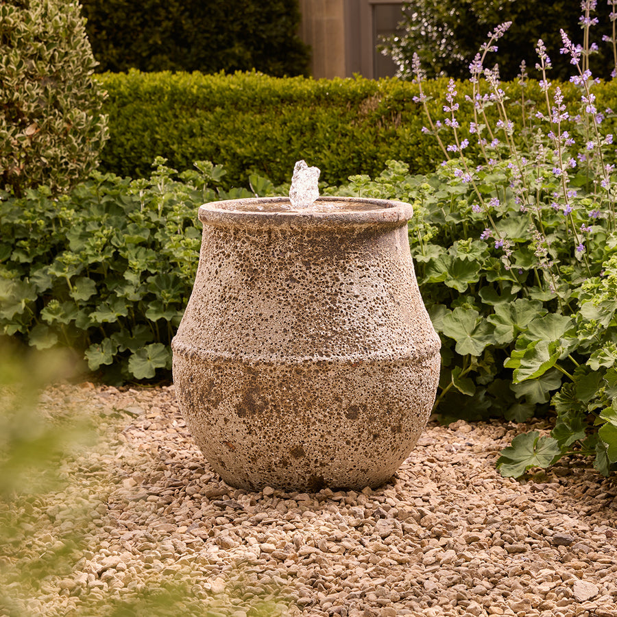 Decorative tall stone water fountain in a garden setting with plants and gravel.