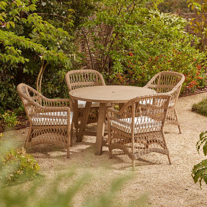 A round outdoor garden dining set featuring a wooden table and four wicker-style armchairs with white seat cushions on a gravel patio