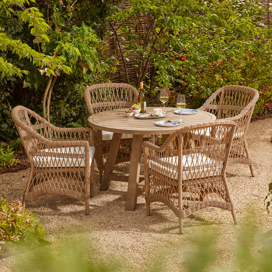 A four-seater round outdoor dining set with a wooden table and wicker-style armchairs, each with white seat cushions, set on a gravel patio surrounded by lush greenery in a garden setting.
