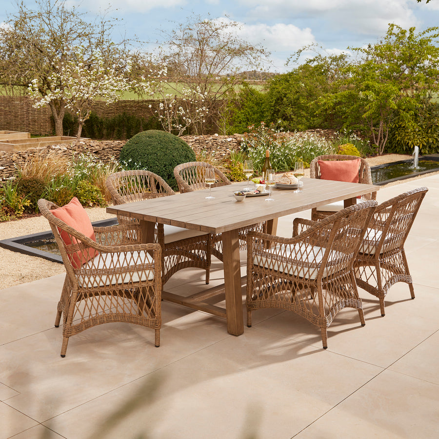 All weather open weave rattan garden dining for six people. Features a handcrafted acacia wooden table and six armchairs with comfortable seat pads, styled on a stone terrace.