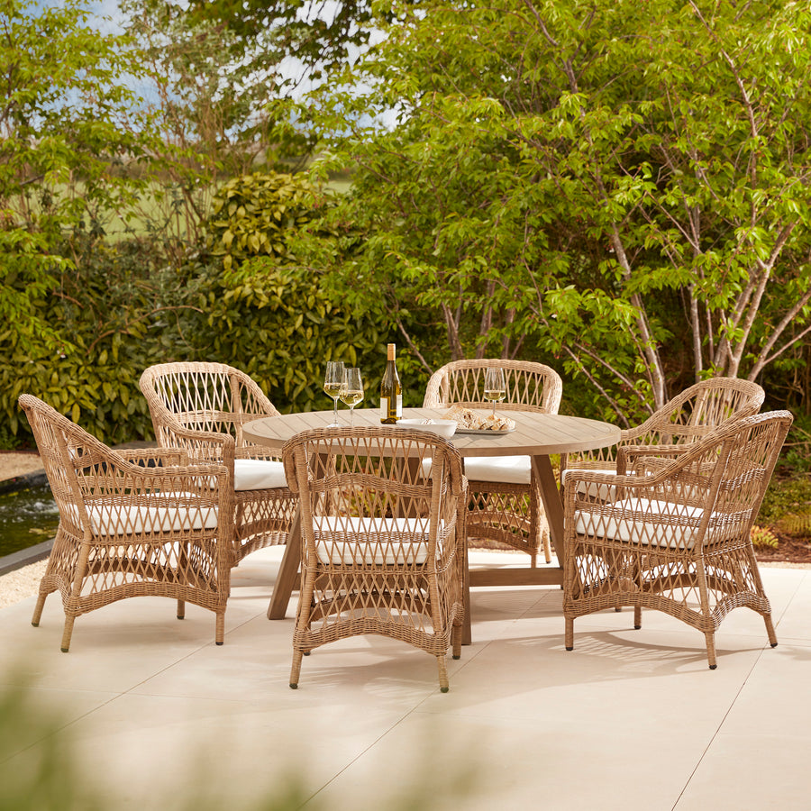 A six-seater round outdoor dining set featuring a wooden table and wicker-style armchairs with white seat cushions, set on a gravel patio and surrounded by lush green garden foliage.
