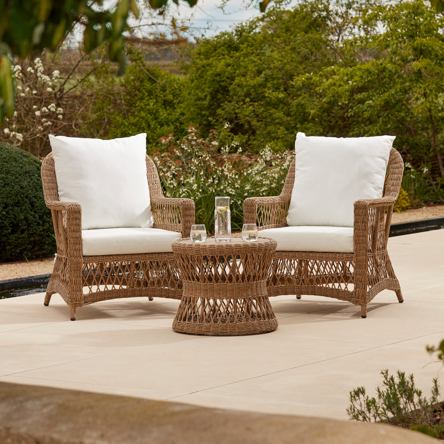 A two-seater round side table and chair set featuring a wooden and open weave table and wicker-style armchairs. Set on a grey terrace 