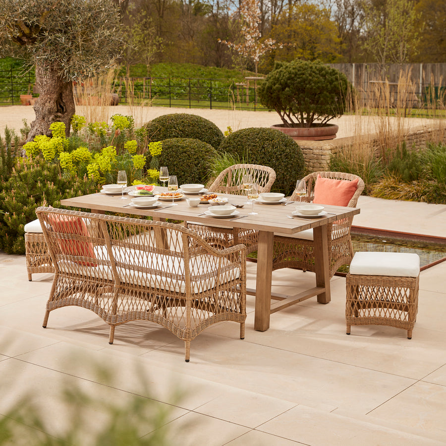 All weather open weave rattan garden dining set featuring a handcrafted acacia wooden table, woven sofa seating perfect for families with small children, and armchairs with deep, cushioned pads, styled on a stone terrace amidst sculpted greenery.