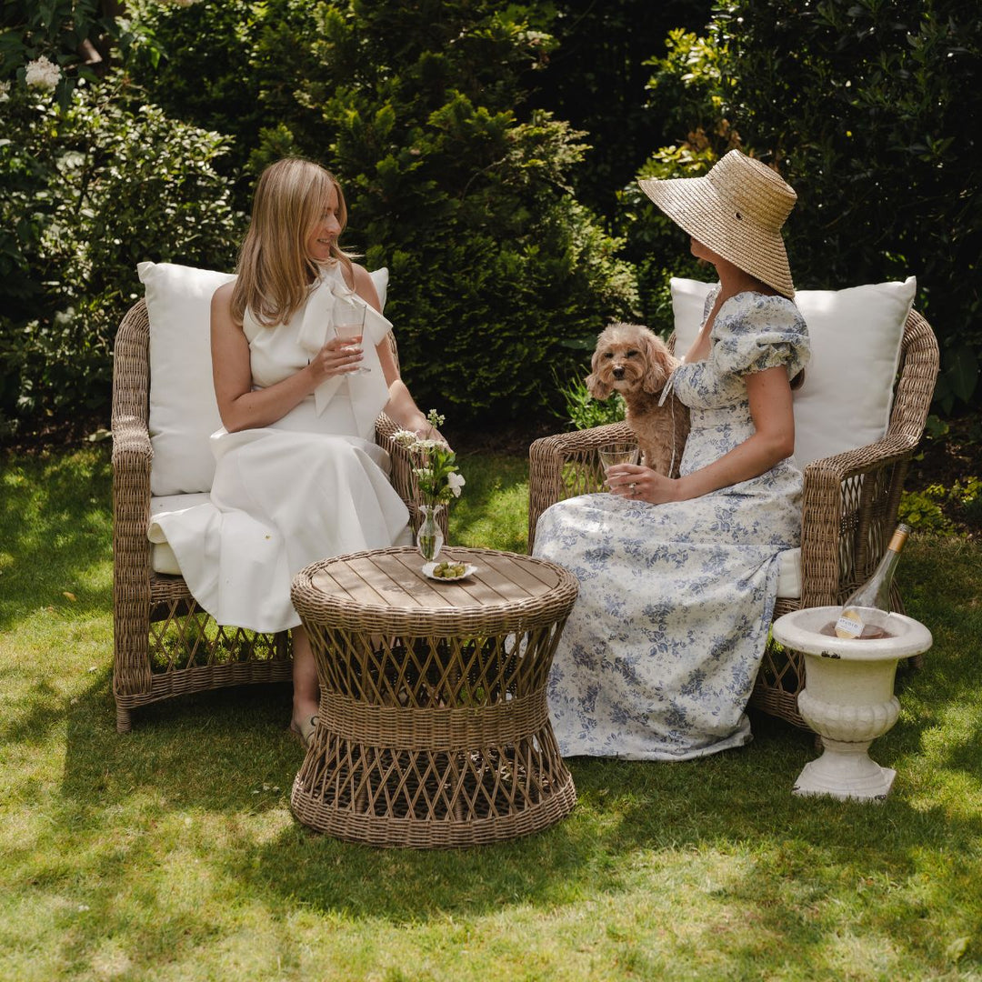 Two women sitting in wicker chairs with a small dog on a grassy lawn.