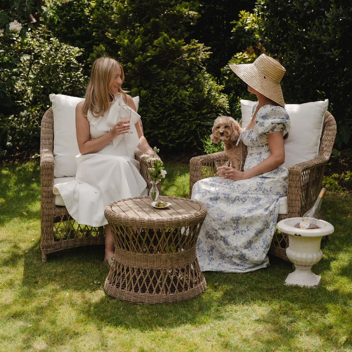Two women sitting in wicker chairs with a small dog on a grassy lawn.