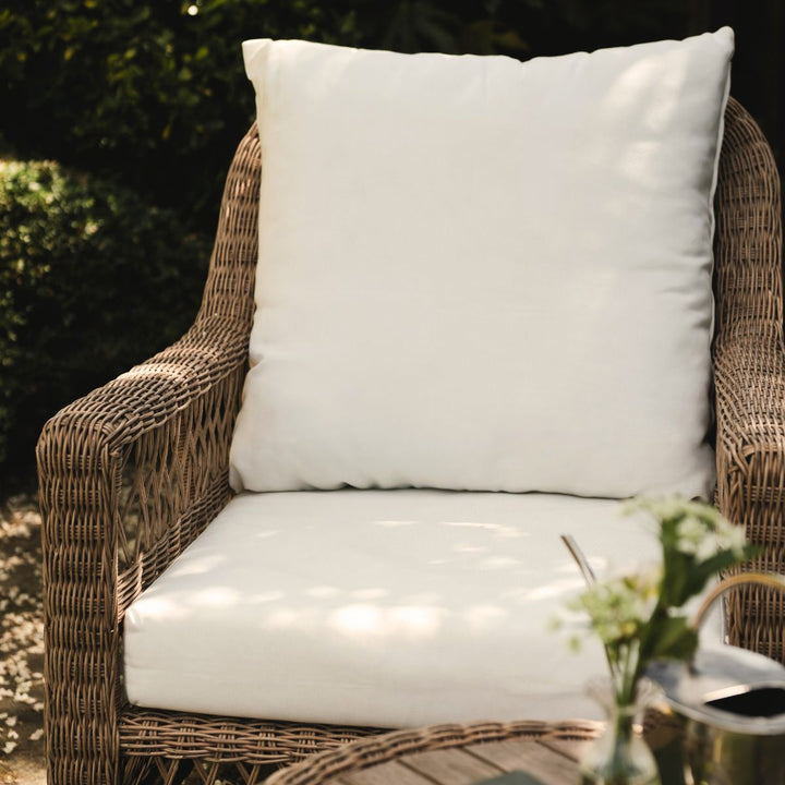 Wicker chair with plump white cushion and small table with flowers in a garden setting