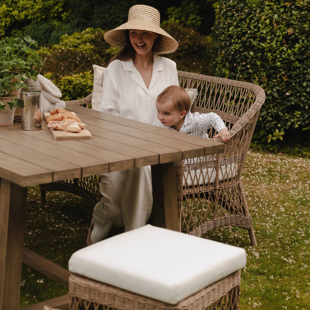 Woman and child sitting on a spacious garden dining sofa behind a solid wood, farmhouse table outdoors.