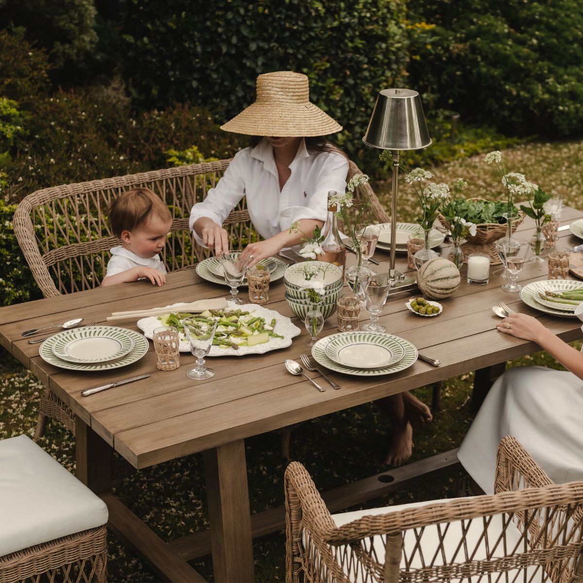 Woman and child sitting at a wooden outdoor table with a meal, surrounded by garden greenery.