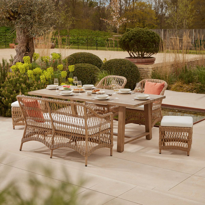All weather open weave rattan garden dining set featuring a handcrafted acacia wooden table, woven sofa seating perfect for families with small children, and armchairs with deep, cushioned pads, styled on a stone terrace amidst sculpted greenery.